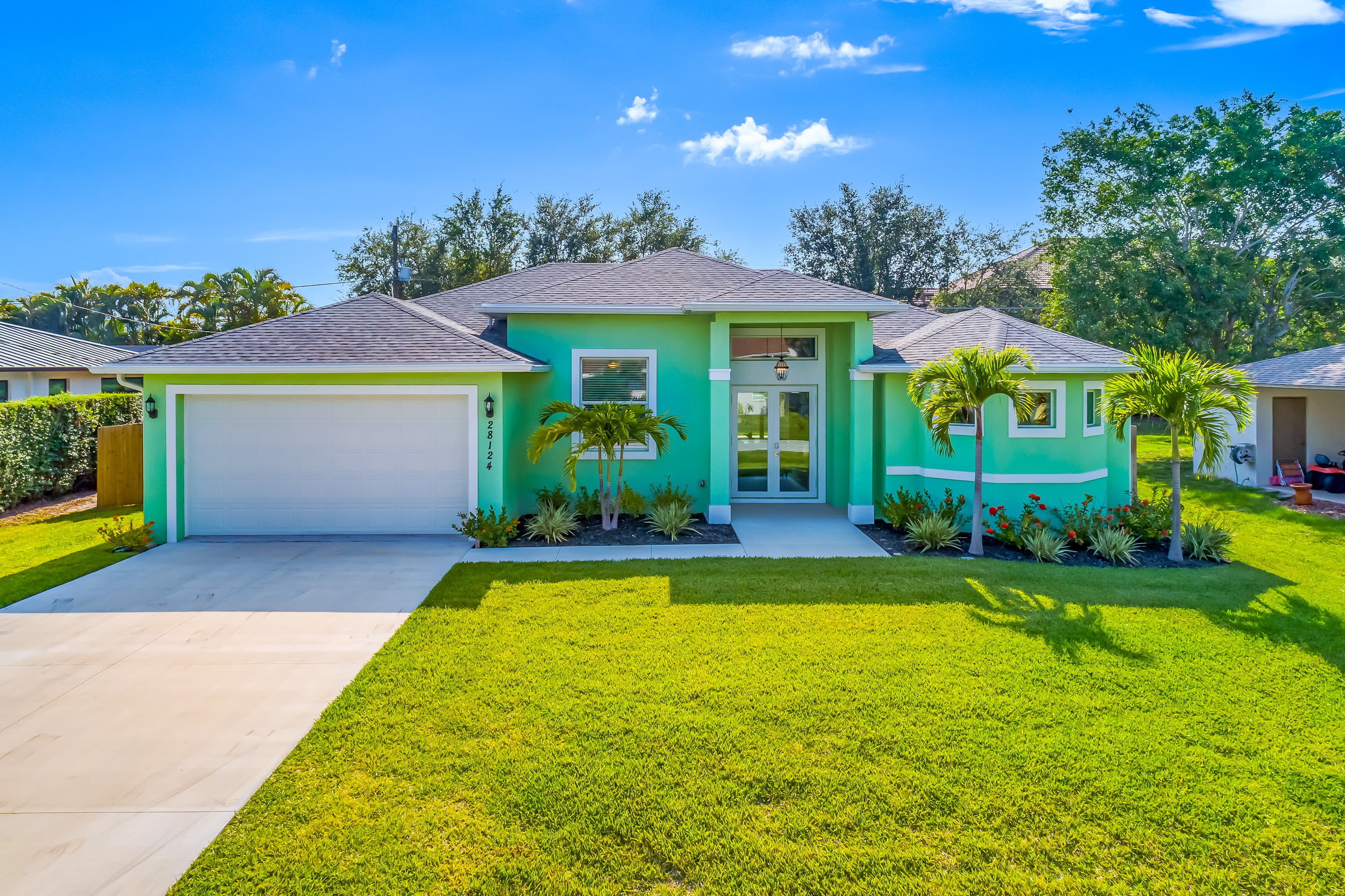 Front view of a single-story sea green house with white double doors, two palm trees in front, garage, and green lawn under sunny blue sky