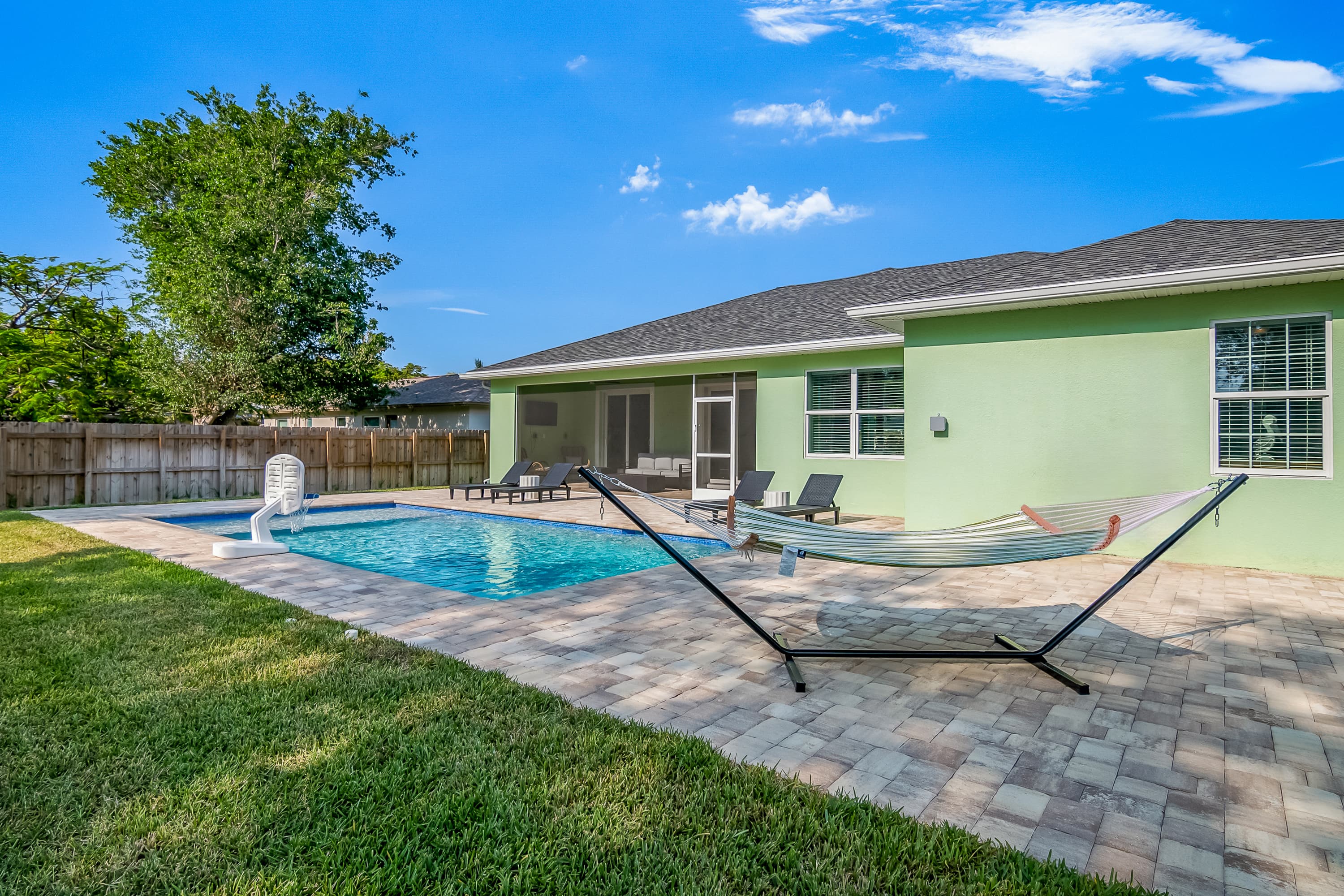 Swimming pool with basketball hoop, stone patio, hammock on stand, and light green house with screen porch
