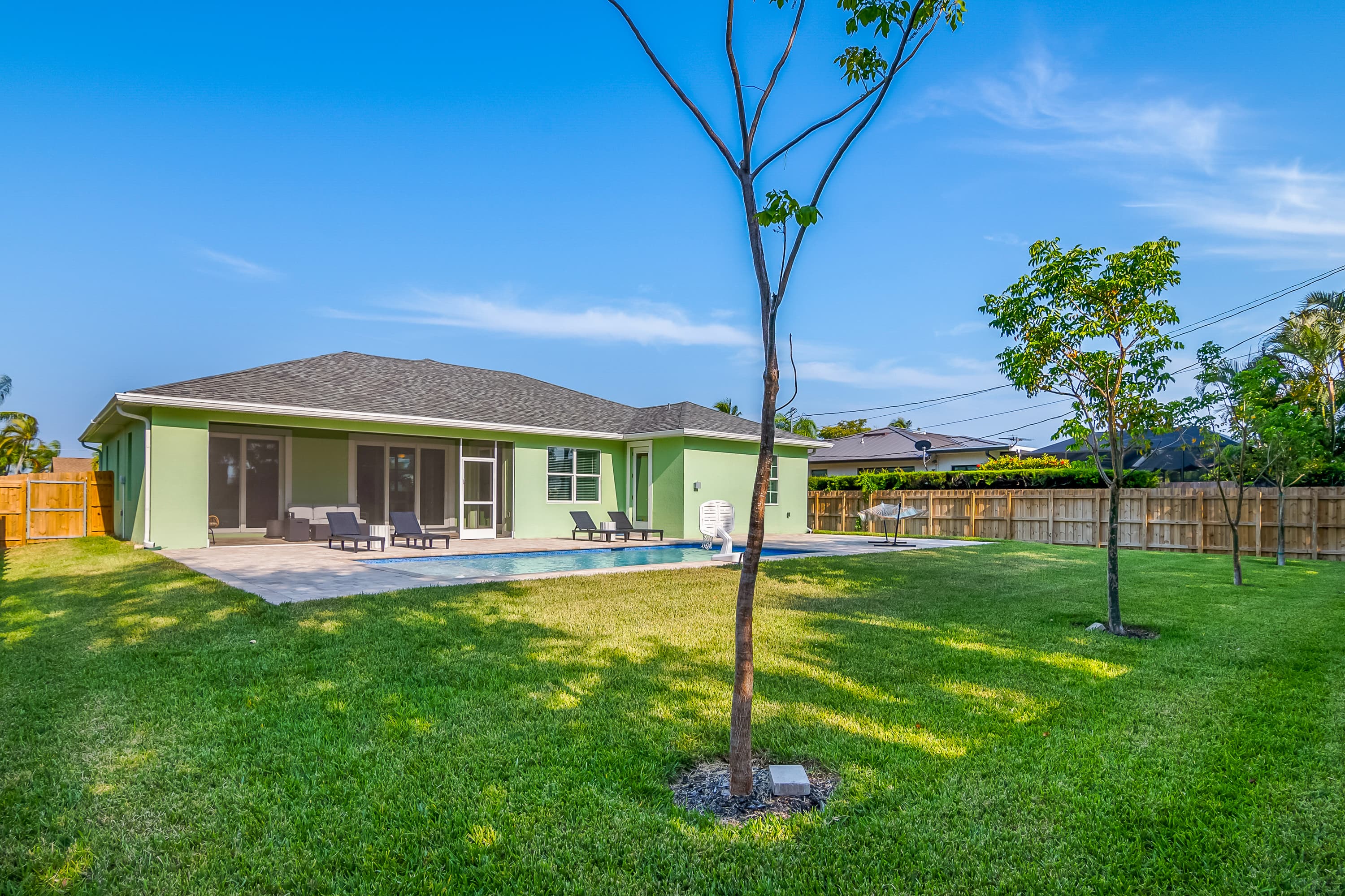 Blue rectangular swimming pool with stone decking, portable basketball hoop, and green grass beyond fenced backyard